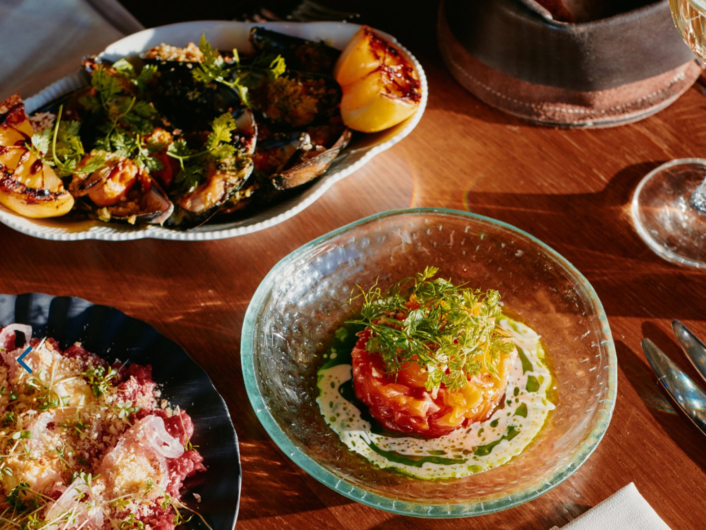Utsnitt av forsiden til oslobukta.no: matbildet fra toppsaken, med blåskjell, tartar i glasskål og et hvitvinsglass på et solbelyst restaurantbord.